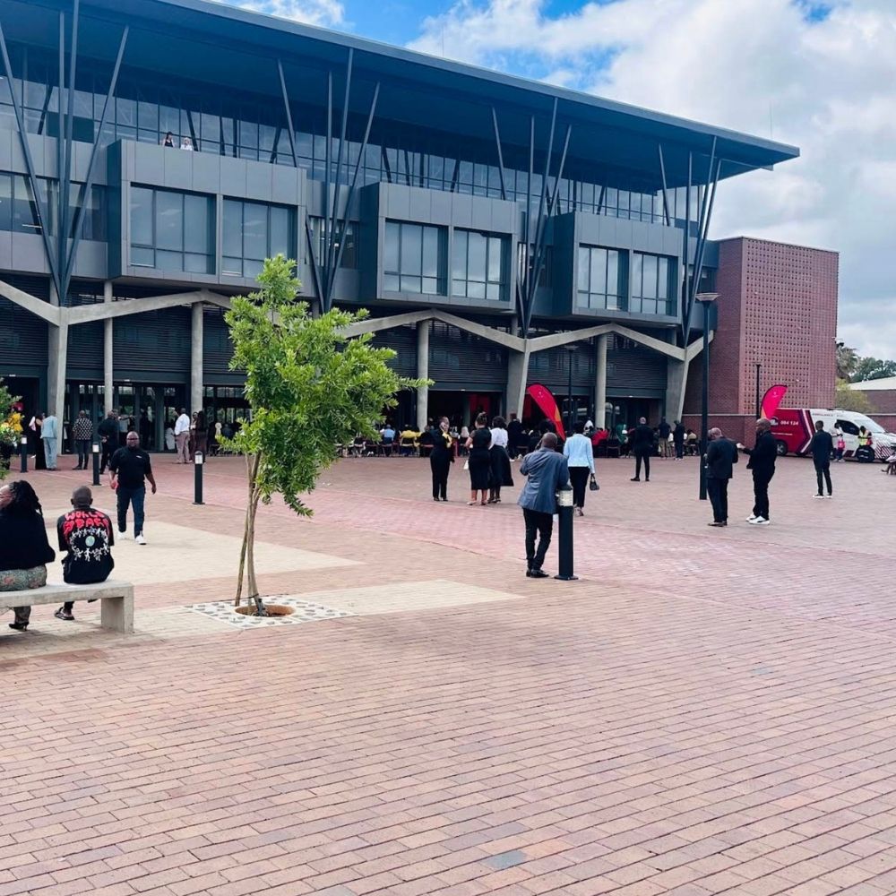 Plaza Gathering at Sol Plaatje University People gather in the brick plaza outside a modern glass building with an angular canopy roof at Sol Plaatje University.